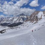 Image of snowy mountains on a blue sky day. Two hikers can be seen in the background hiking through the snow.