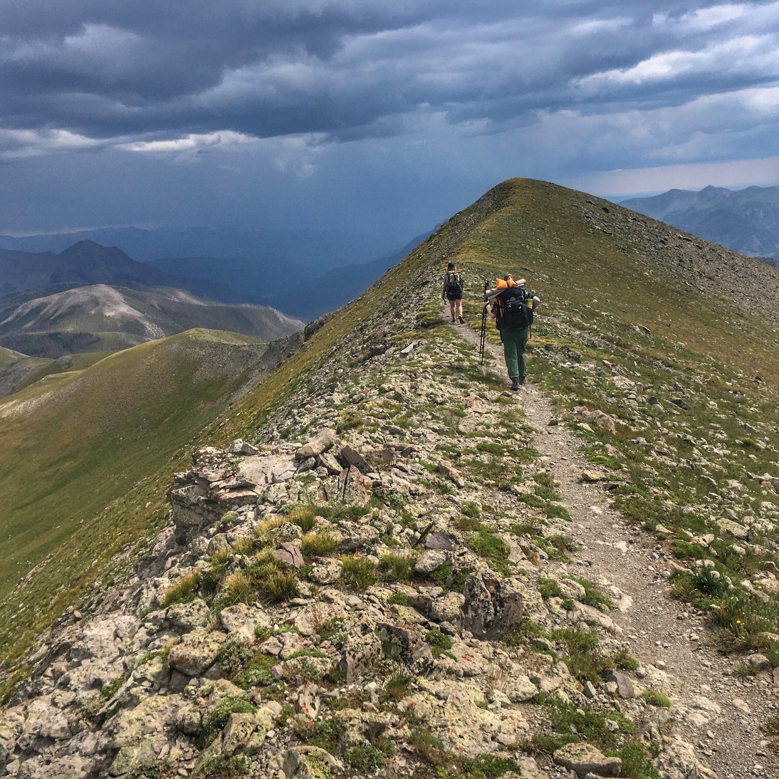 Two hikers on a mountain with dramatic storm clouds in the background.