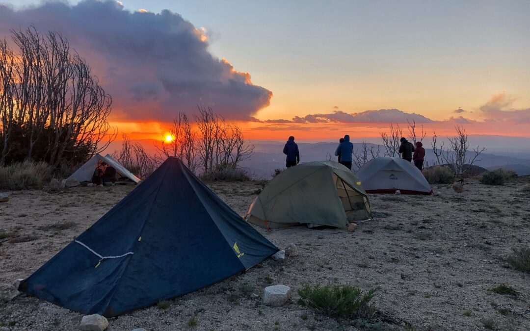 A group of hikers watching the sunset with their tents set up on a mountain.