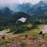 Three hikers climbing a mountain with a lake in the background. The landscape is green and there is mist in the peaks seen in the background.